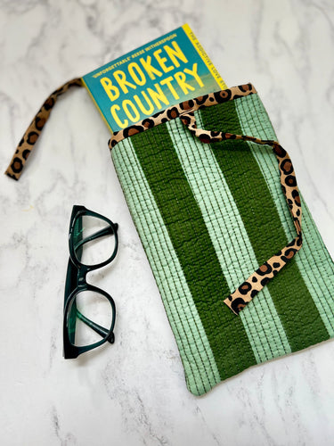 A green striped quilted book pouch with leopard-print ties, shown on a white marble surface with a book and eyeglasses as props.